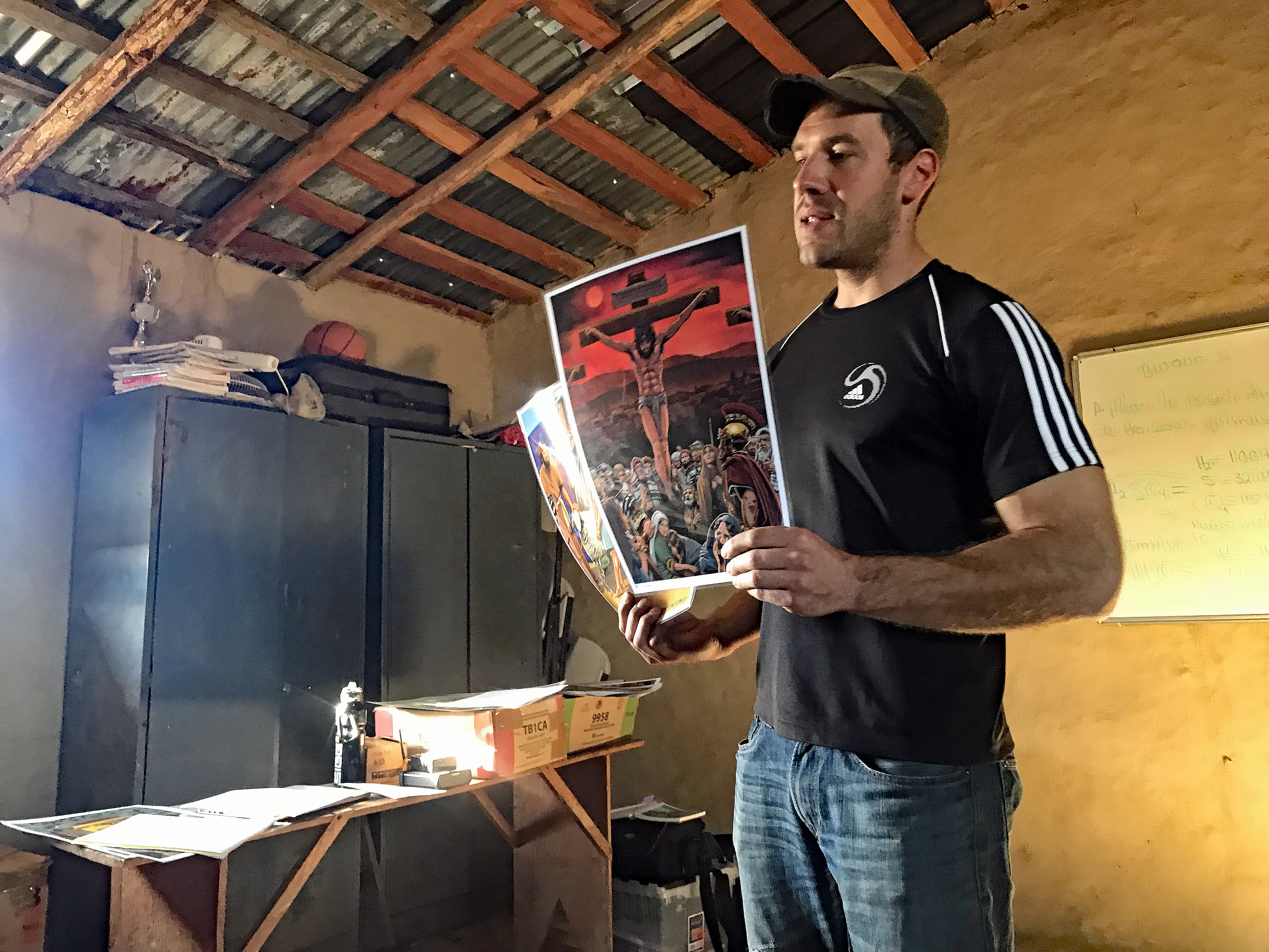 Our coworker Pete, holding lesson notes and an illustration of Christ on the Cross, teaching inside a school room in Las Moras