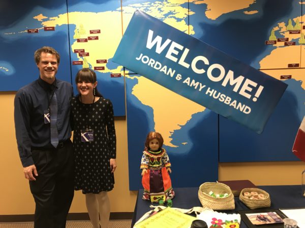 Jordan and Amy standing in front of the missions world map at Crossroads Bible Church in Bellevue, WA. To one side appears a table display including - among other details - a doll in traditional Nahuatl dress and various Mexican candies for visitors to sample.