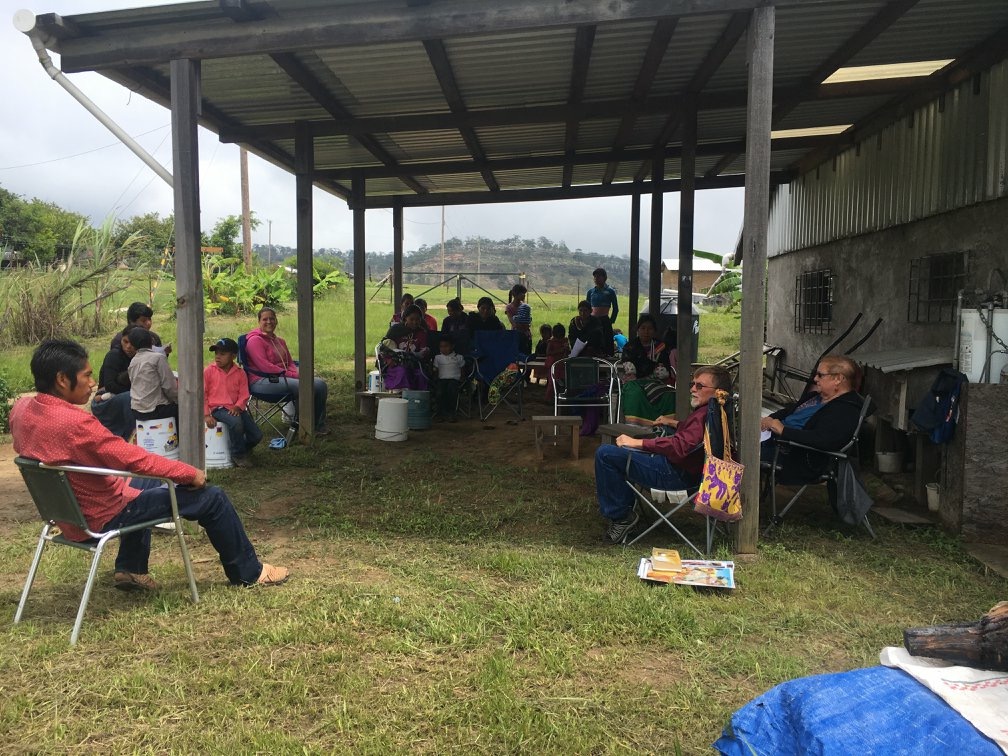 A number of people sitting in camping chairs, in or near the shade of an open carport, with grass below.