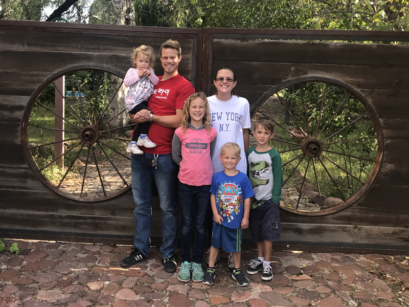 Lily, Jordan, Elayne, Amy, Joel, and Titus in front of a double-doored wooden gate with large wagon wheels inset into the panels.